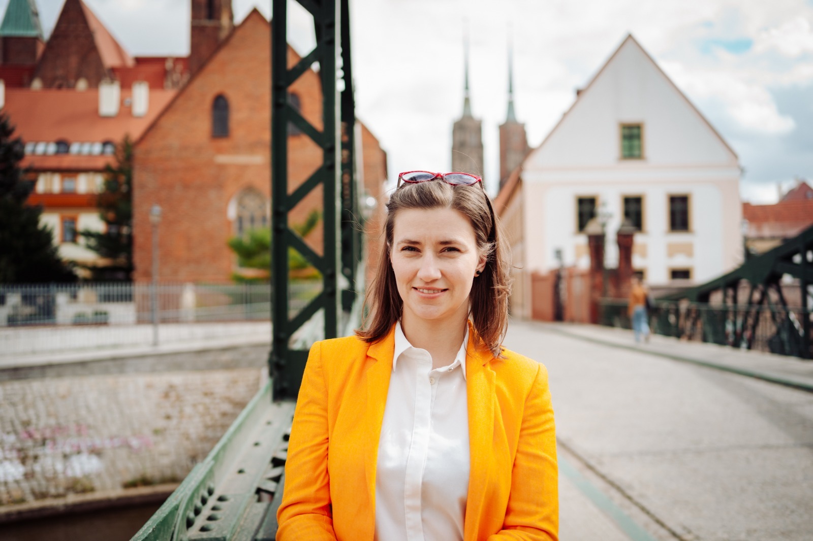 Osoba w pomarańczowej marynarce stojąca na moście, w tle zabytkowa zabudowa i wieże kościelne